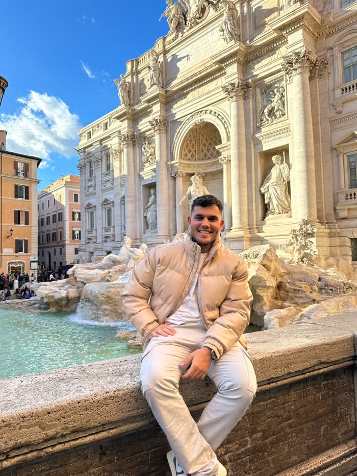Alexandre sentado em frente à Fontana di Trevi, sob o céu azul